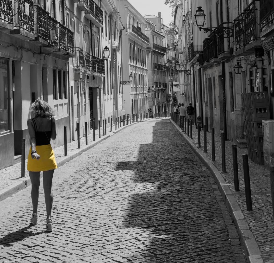A black and white picture of a woman crossing a paved street half obscured by shadow of the buildings on the right. The woman's skirt is the only element in color, a bright yellow.