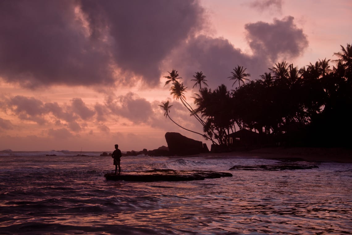A purply sunset over turtle beach, with a fisherman standing in the foreground and palm trees in the back